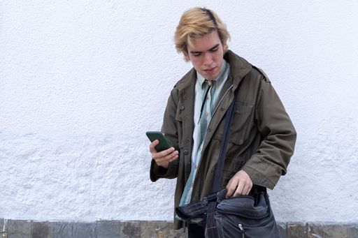 A man holding his phone and taking something out from his bag while standing in front of a white wall