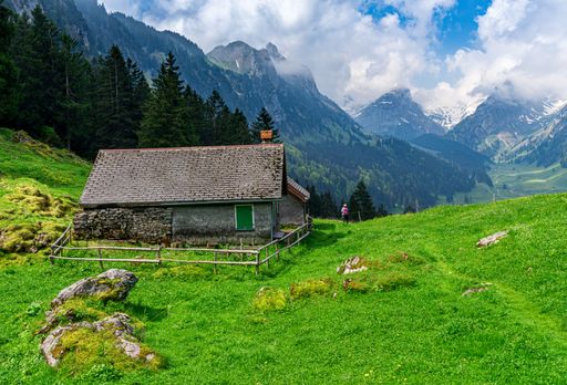 An old rustic cabin is nestled in the majestic mountains, surrounded by lush green grassy hills