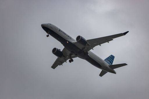 A JetBlue Airbus A220 aircraft flying through the sky on a grey cloudy day