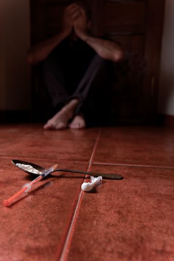 man lying on the floor in the dark with his hands on his head desolate from drugs in the foreground syringe with heroin and spoon