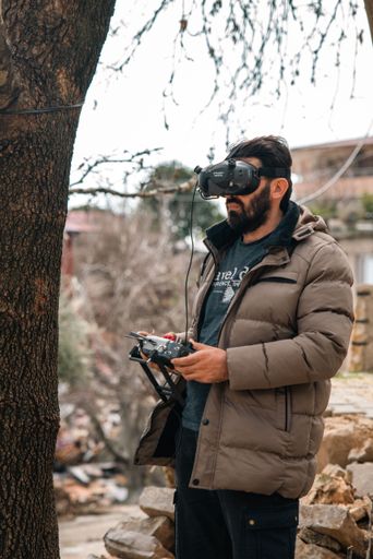 A young male wearing virtual reality glasses and a hooded jacket standing next to a tall tree