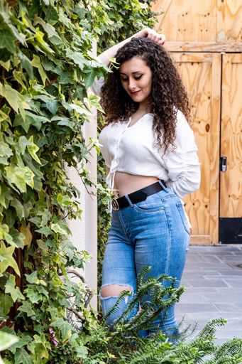 vertical shot of a beautiful young woman with long curly brown hair, next to a wall of ivy full of leaves looking down, in a white t-shirt and jeans