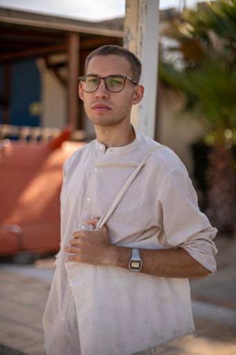 a young Caucasian man in a collarless shirt and dress pants, leaning on a stick and holding a shopping bag