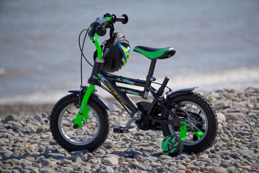 Colorful bike for kids with protective helmet hanged on the handlebars and mounted training wheels parked on pebble beach on sunny day.