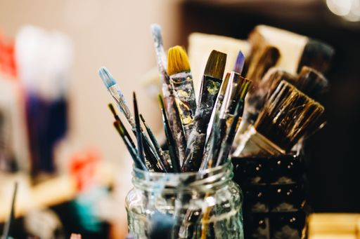A closeup of paintbrushes and acrylic paint in a glass jar with a blurry background
