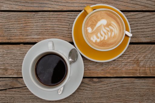 A top-view of a cup of cappuccino with latte art and a coffee cup on a saucer filled with black coffee on a wooden table