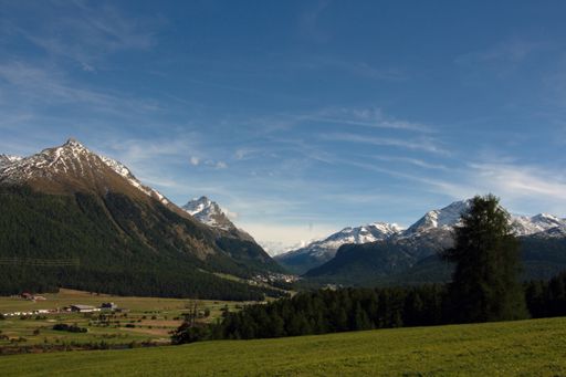 A scenic view of a green field on a sunny day surrounded by high mountains