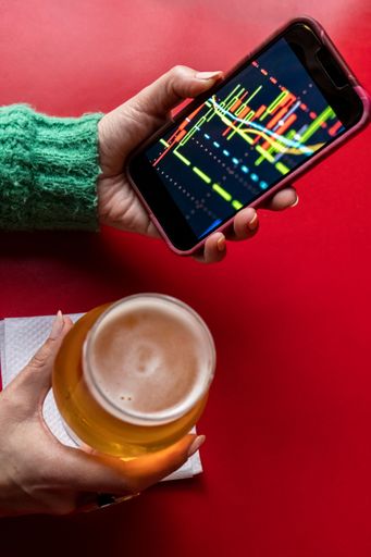 A top view of female hand holding a phone with graphs on screen and a glass of beer on a red backdrop