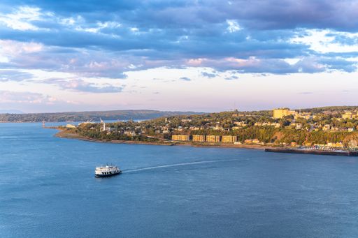A drone Scenic view of a boat in calm blue ocean at sunset with greenery and buildings in the background in Quebec City, Canada