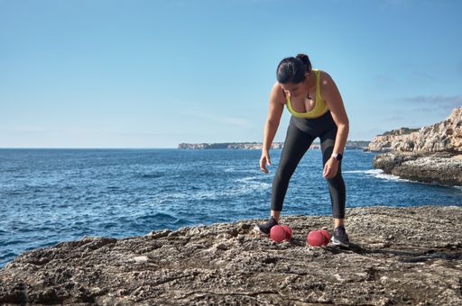 A Hispanic female in a sports outfit working out and doing exercises on the beach near rocky cliffs