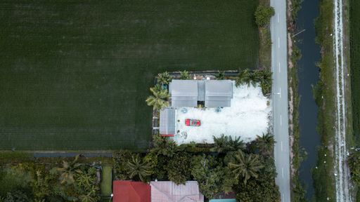 An aerial top view of a rural house with a car in a green field with palm trees