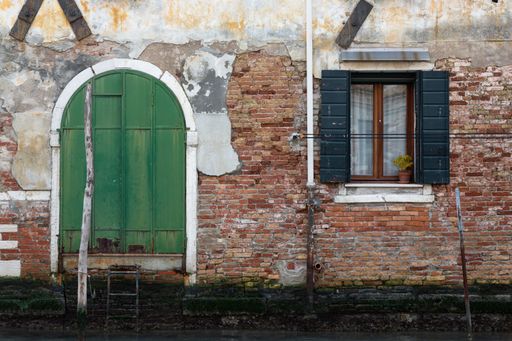 An old brick building with a green arch-shaped door and black window shutters by the canal in Venice