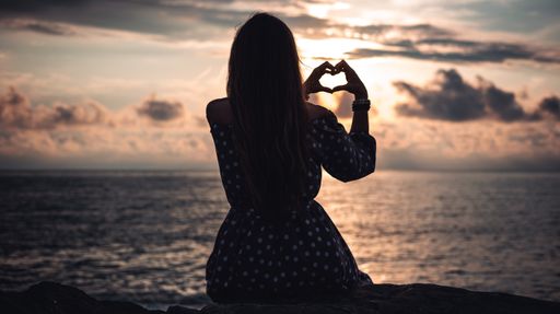 A silhouette of a female sitting on a beach showing a heart gesture at the golden hour