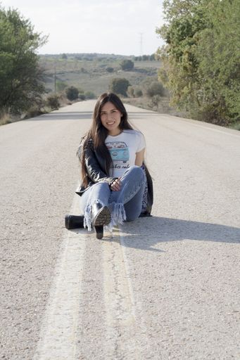 A  young Latina female wearing jeans, bodysuit and leather jacket and posing on an asphalt road