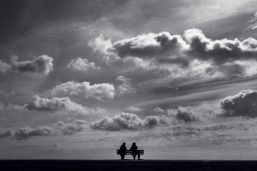 A romantic silhouette of a couple sitting on a bench at a field under a cloudy sky in grayscal
