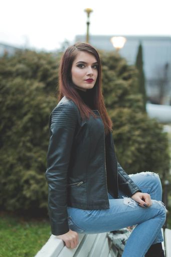 A vertical shot of a young brunette female in a leather coat posing on a bench