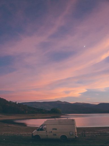 A vertical shot of a van by the sea surrounded by high mountains under the sunset sky