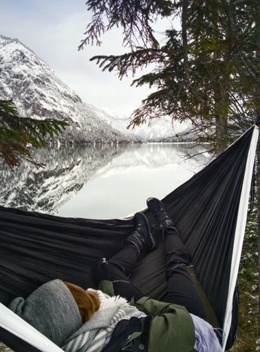 A vertical shot of a person resting in a hammock by a lake surrounded by beautiful snow-covered mountains