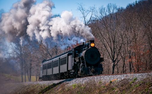 A restored steam passenger train moving slowly blowing lots of black smoke and white steam on a sunny day.
