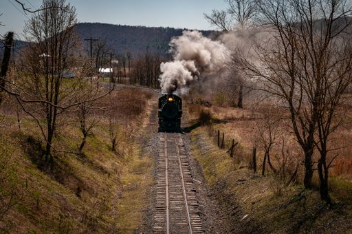 A restored steam passenger train moving slowly blowing lots of black smoke and white steam on a sunny day.