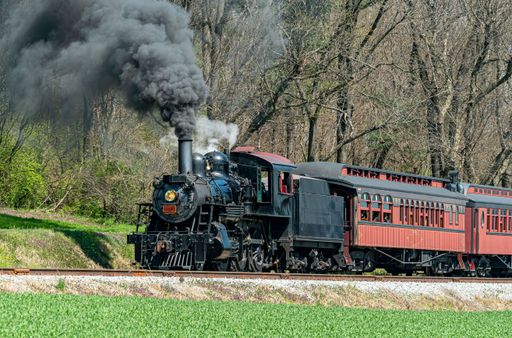A restored steam passenger train moving slowly blowing lots of black smoke and white steam on a sunny day.