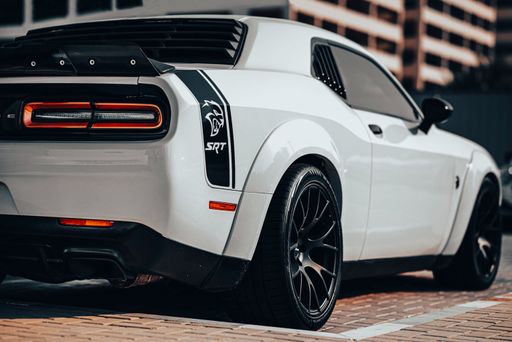 A white Dodge Hellcat SRT car is parked in front of a large building, illuminated by the sun