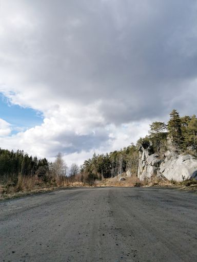 A vertical shot of an empty road in Ranvika, Norway, next to cliffs and trees under huge clouds in the sky