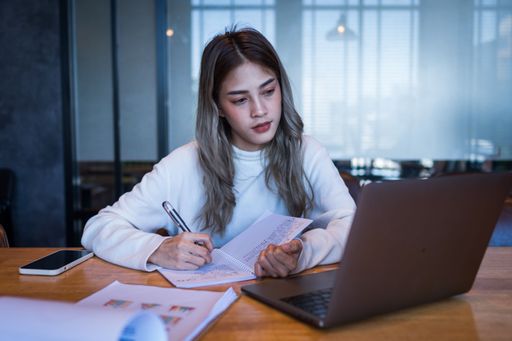 A female seated at a desk, looking at her laptop while taking notes