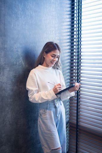 A female is seen using a digital tablet with a pen while leaning against a brick wall