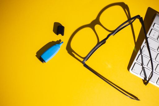 A top view of glasses, keyboard and a blue marker on a yellow work table