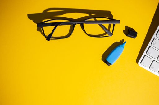 A top view of glasses, keyboard and a blue marker on a yellow work table