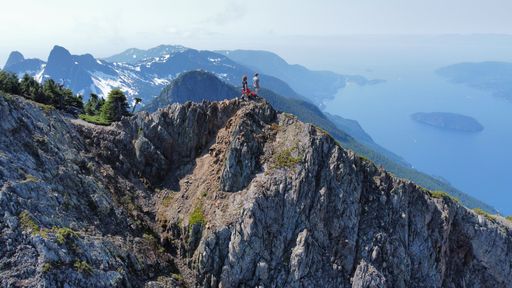 Two male adults standing atop a jagged rocky mountain overlooking a tranquil lake below
