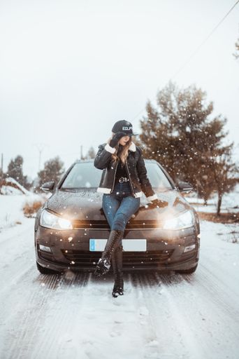 A vertical shot of a Hispanic sexy, cocky girl leaning on a background of a black car with lights on