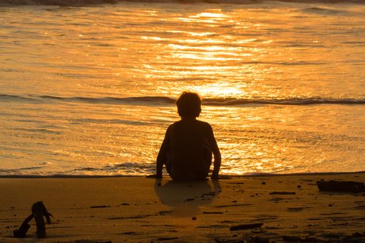 A silhouette of a boy admiring the golden sky reflection on the water sitting alone on a sandy shore