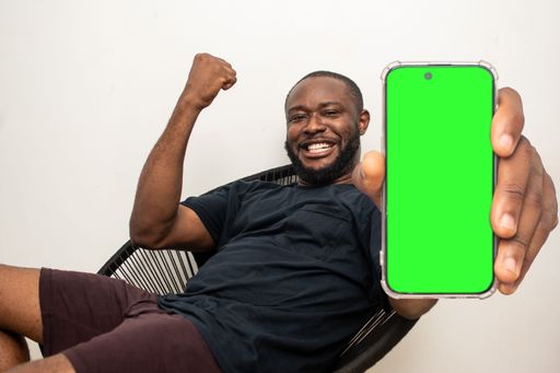 A young African man holding his phone gesturing a win while sitting in a chair, with copy space