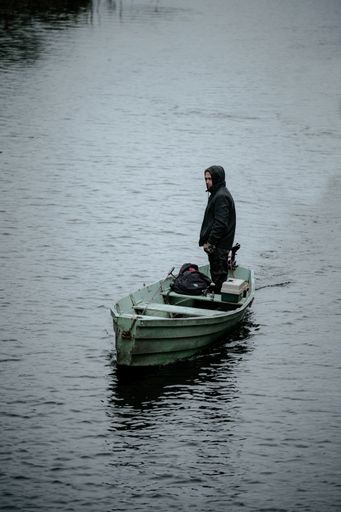 A young man in a warm winter outfit standing on small rowboat in the middle of a lake on a gloomy day