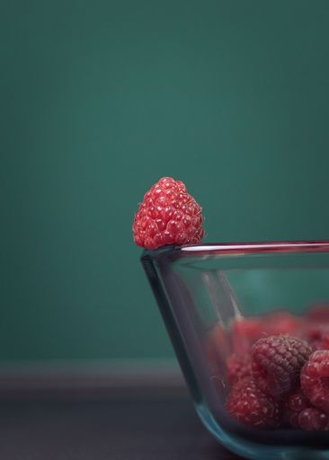 A glass bowl containing ripe raspberries against a green background