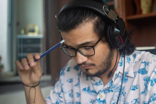 A Hispanic man with headphones, pressing on his forehead with a pencil while working at home
