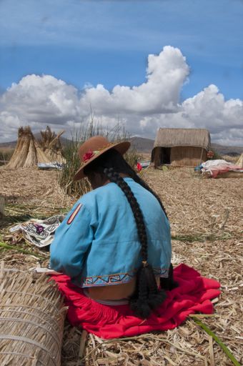 A vertical shot of a woman wearing blue and red with a hat showing long braided hair from behind, sitting on a beach at a village under cloudy sky