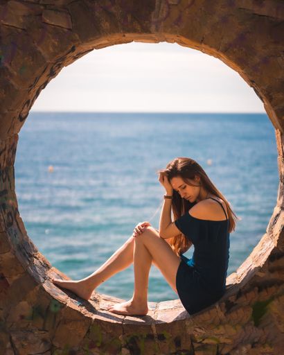 A young attractive woman from Spain in a blue dress sitting in the stone round window by the sea