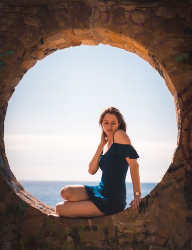 A young attractive woman from Spain in a blue dress sitting in the stone round window by the sea