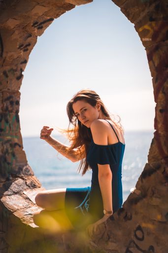 A young attractive woman from Spain in a blue dress sitting in the stone round window by the sea