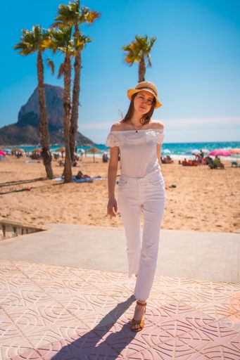 An attractive young woman from Spain dressed in white with a straw hat walking along the beach of Calpe, Valencia, Spain.