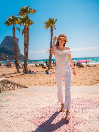 An attractive young woman from Spain dressed in white with a straw hat walking along the beach of Calpe, Valencia, Spain.
