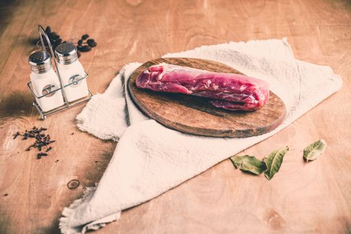 A piece of raw pork tenderloin on a chopping board with seasonings on the table