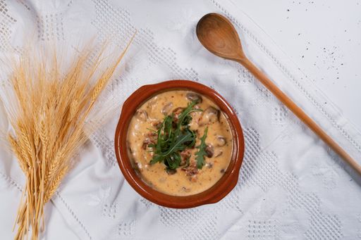 A top view of soup with mushrooms and greens on a white tablecloth with a wooden spoon and wheat