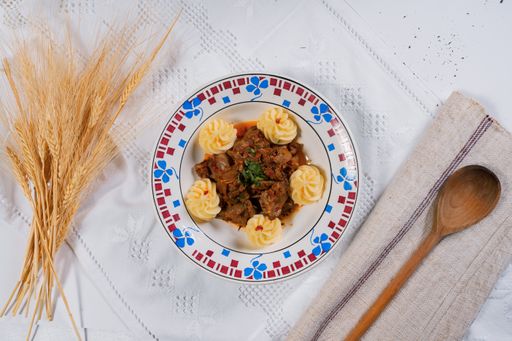 A top view of a beef dish with dumplings on a white tablecloth with a wooden spoon and wheat sheaves