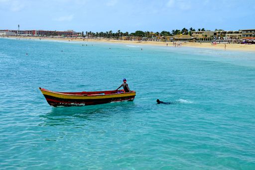 Boat in sea carrying a man, with another man swimming behind it, near beach at Santa Maria, Sal Island, Cape Verde.