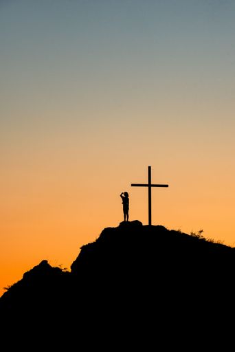 A silhouette of a person standing near a cross on a hill during the sunset