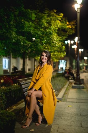A vertical shot of a young blonde female model in a fashionable yellow coat posing on a bench